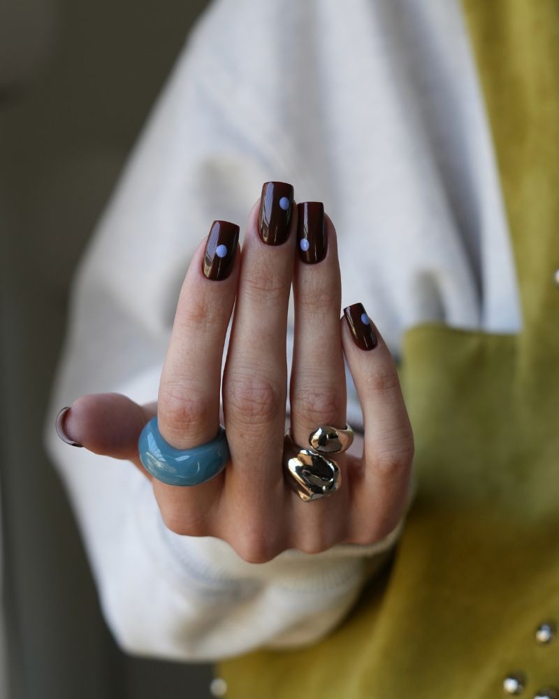 deep oxblood wine brown short square nails with two small lavender blue polka dots near the tip blue ceramic ring gold sculptural ring white mustard jacket