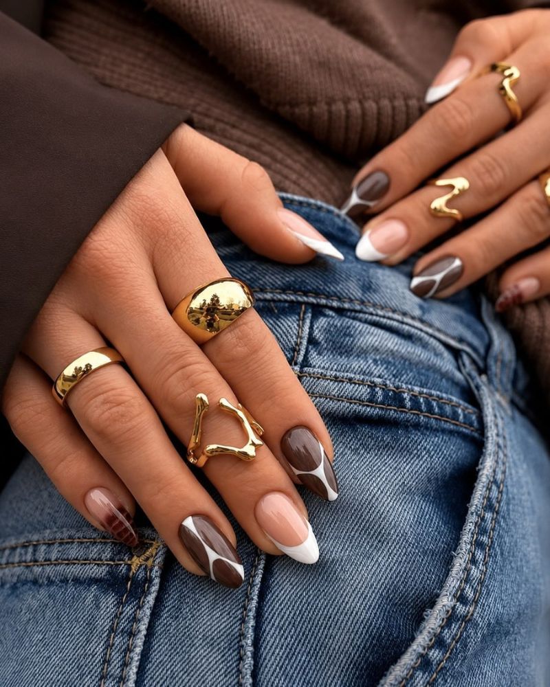 short almond nails mixed set warm brown solid nude nails with abstract brown white cow blob pattern and white french tips gold rings blue denim brown jacket