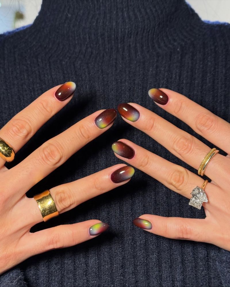 short oval autumn aura nails dark wine chocolate center bleeding into orange amber yellow green edges both hands on chest navy turtleneck multiple gold rings