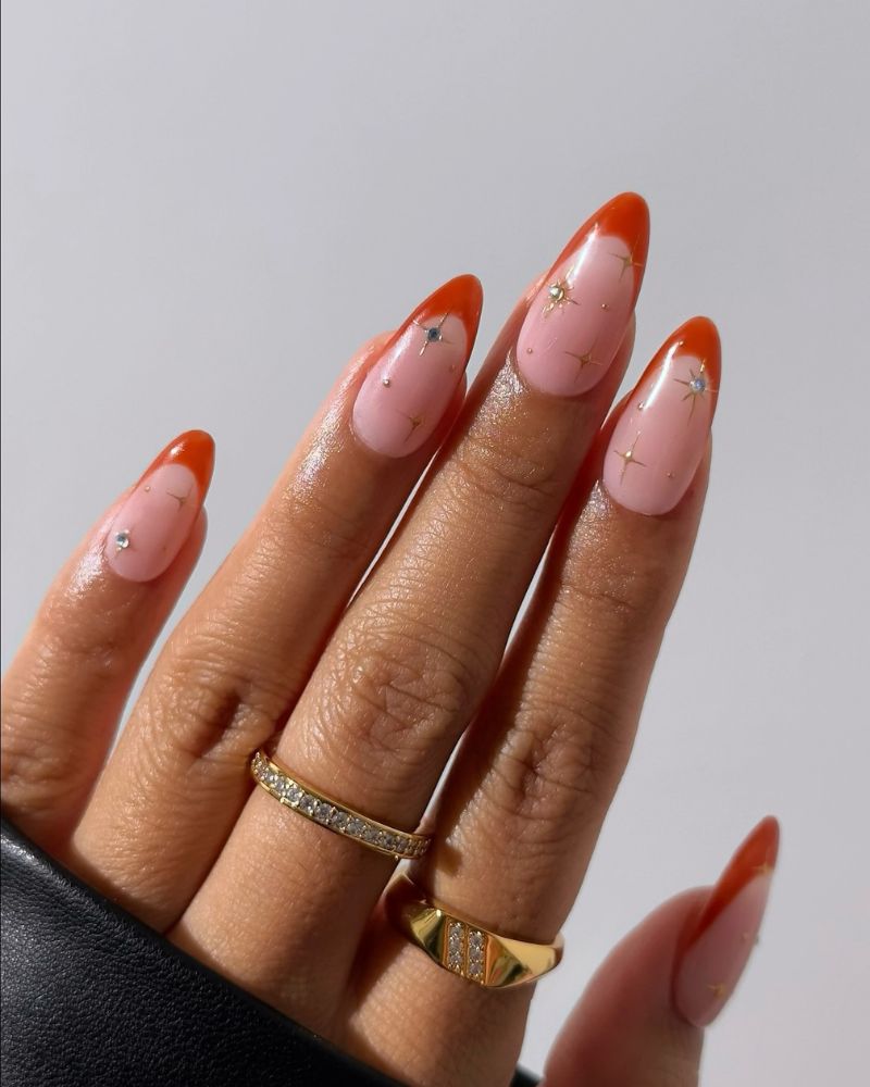 burnt orange french tip on sheer pink almond nails with scattered gold star gems